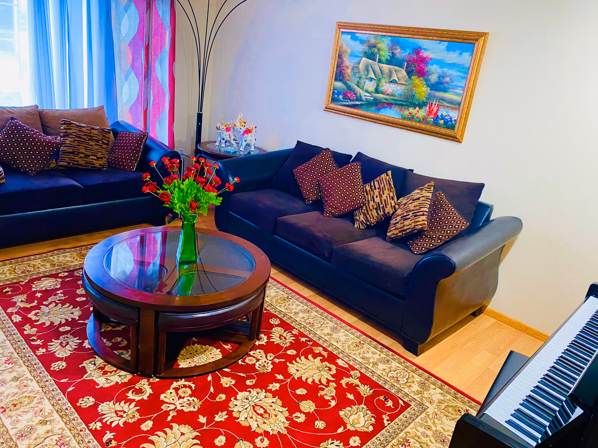 A cozy living room with two dark blue sofas adorned with patterned cushions, a round wooden coffee table with a glass top and a vase of red flowers, a colorful floral area rug, a floor lamp, a side table with decorative items, a framed painting of a cottage and garden on the wall, and a piano keyboard visible on the right side.