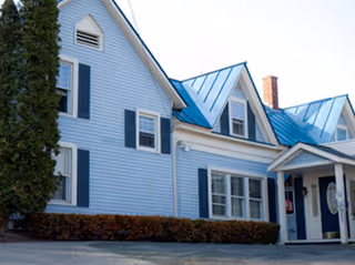 Exterior view of a light blue building with a blue metal roof, multiple windows with black shutters, a covered entrance with white pillars, and a tall evergreen tree on the left side.