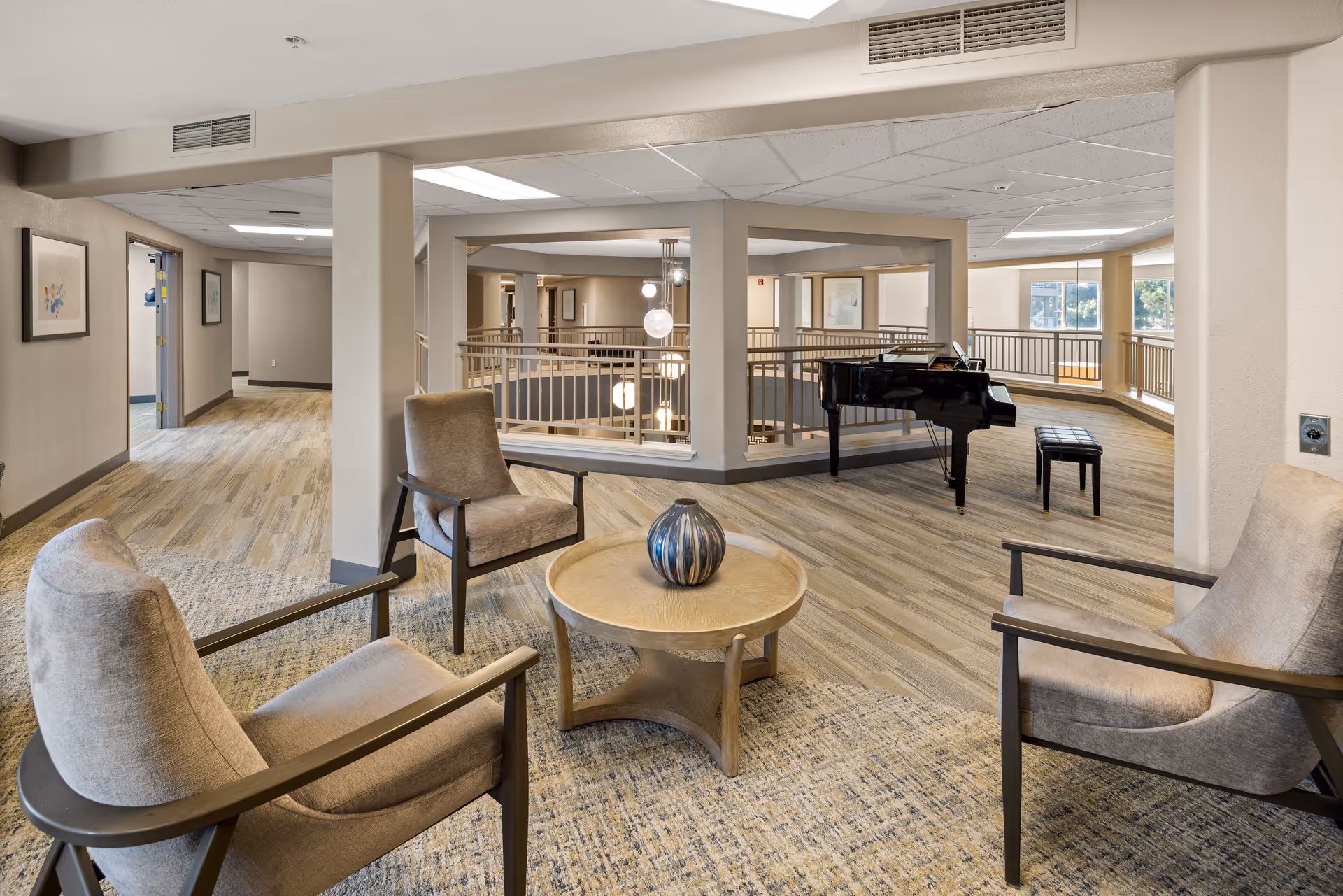 A spacious senior living community lounge area with three beige cushioned chairs arranged around a round wooden table with a decorative vase. In the background, there is a black grand piano with a matching bench near large windows letting in natural light. The floor is a combination of carpet and wood-like flooring, and the walls are painted in neutral tones. The area overlooks a lower level with a railing and hanging globe light fixtures.