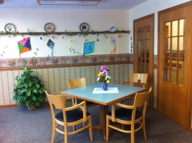 A small dining area with a square table and four wooden chairs with black cushions. A small vase with colorful flowers sits on a white doily in the center of the table. The walls are decorated with a wallpaper border featuring a pattern and a garland with colorful paper kites and plates above it. There is a green potted plant in the corner and two wooden doors with glass panels on the right side.