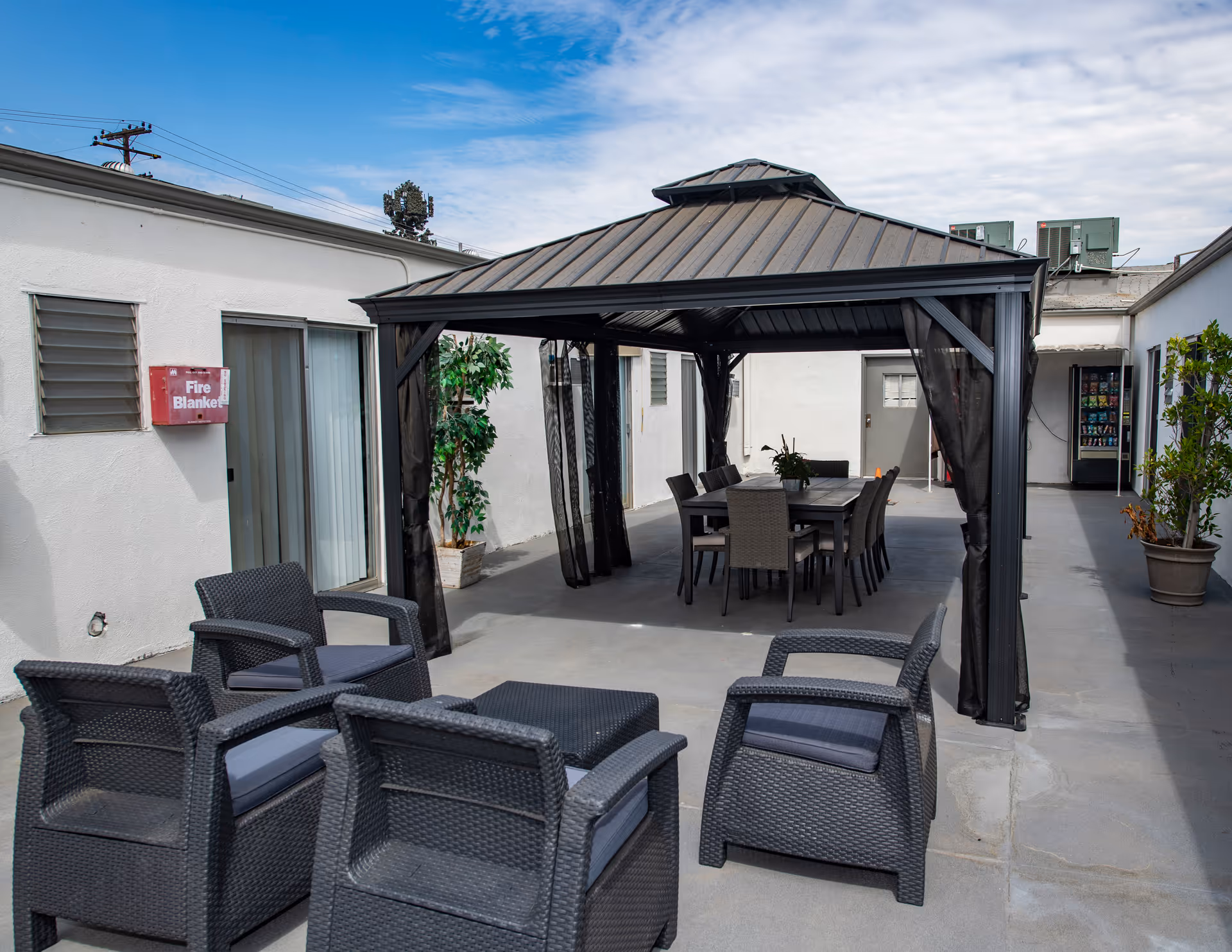 Outdoor patio area at Country Oaks Care Center with a black metal gazebo covering a dining table and chairs. Additional black wicker chairs with cushions are arranged nearby. Potted plants and a vending machine are visible along the white building walls under a partly cloudy sky.