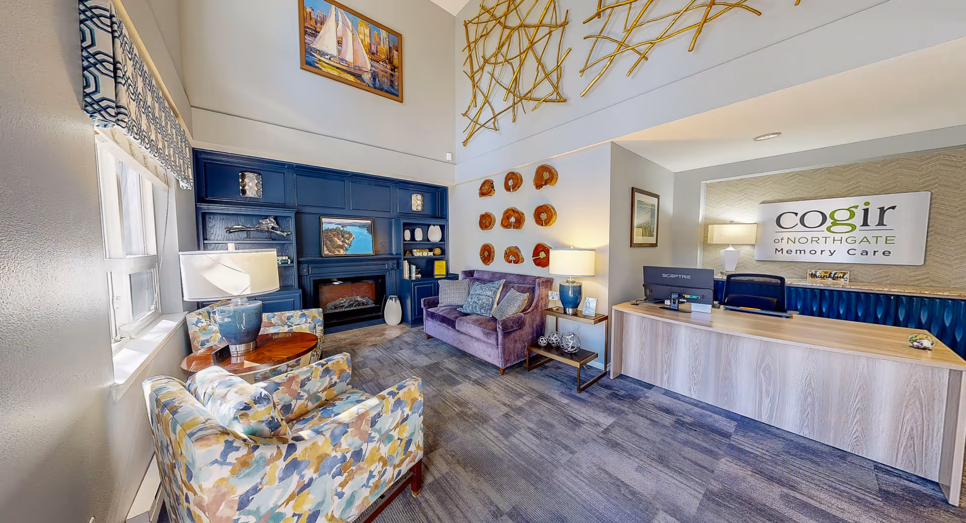A bright and welcoming reception area at Cogir of Northgate Memory Care featuring a wooden reception desk with a computer, a sign on the wall behind the desk, a purple couch, two patterned armchairs, a small round wooden table with a lamp, decorative wall art, and a built-in blue shelving unit with a fireplace and various decorative items.