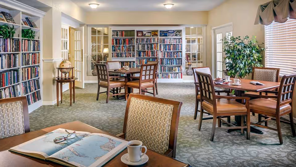 A cozy reading and dining area in an assisted living facility featuring several wooden tables and chairs, bookshelves filled with books, a globe on a small table, a large plant near a window with blinds, and a book and coffee cup on the nearest table.