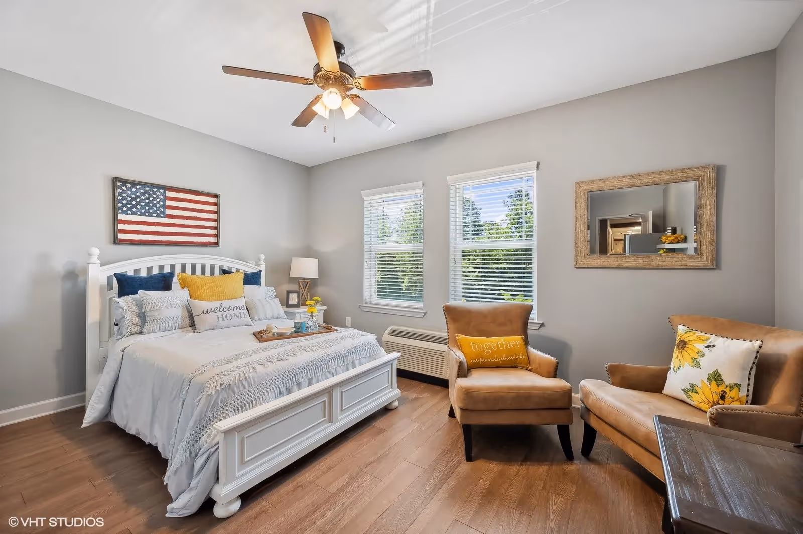 Bright furnished bedroom with a white bed, two tan armchairs, ceiling fan, and American flag wall art.