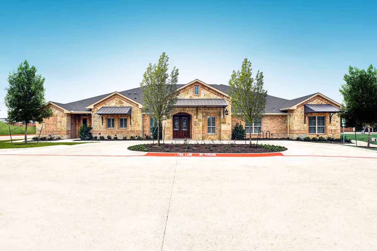 Single-story stone building front with a circular driveway, landscaped island and young trees under a clear sky.