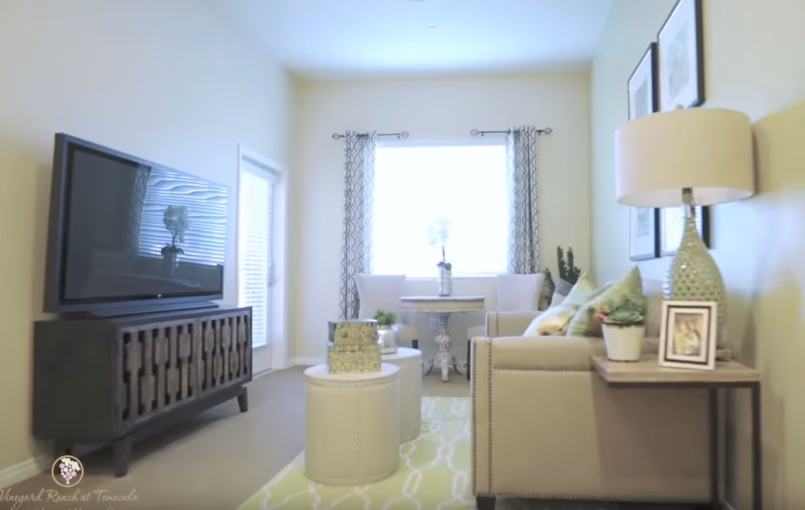 A bright living room with a large window covered by patterned curtains. The room features a beige sofa with green and patterned cushions, a side table with a lamp, a small plant, and a framed photo. Two white ottomans are placed on a light green patterned rug. Across from the sofa is a dark wooden TV stand with a flat-screen TV on top. Two white chairs and a small round table are positioned near the window.