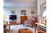 A cozy bedroom in Bethany Home Retirement Center featuring a single bed with a patterned bedspread, a wooden dresser with framed artwork above it, a flat-screen TV on a stand, a side table with a lamp, and a cushioned ottoman in the foreground. The room is softly lit and decorated with additional framed pictures and a vase with flowers on a side table.