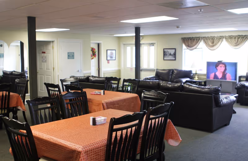 Interior view of a senior living facility common area with several tables covered in orange tablecloths and black chairs arranged around them. In the background, there are black leather sofas facing a television that is turned on. The room has light-colored walls, a carpeted floor, and ceiling lights.