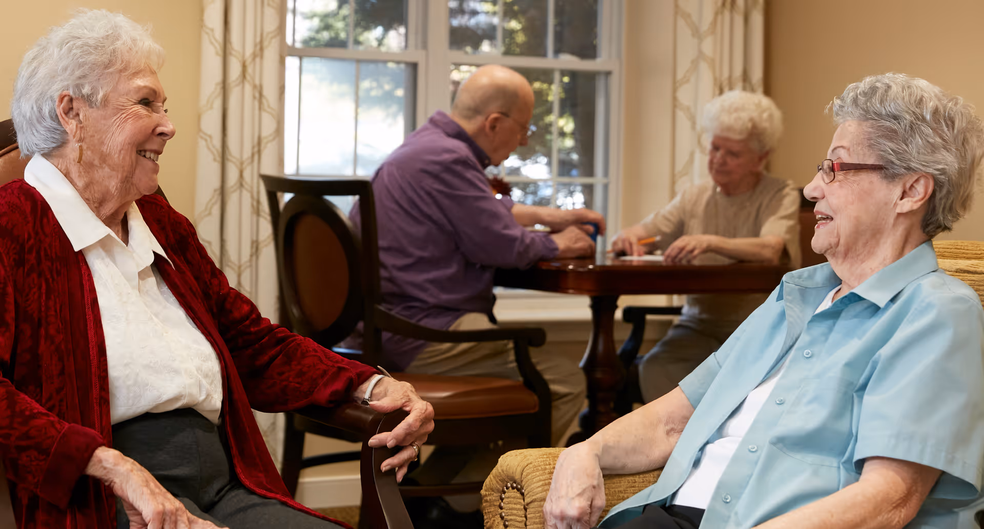 Four elderly individuals in a well-lit room with large windows. Two women are seated in the foreground facing each other and smiling, while a man and another woman are seated at a table in the background engaged in an activity. The room has light-colored walls and curtains with a subtle pattern.