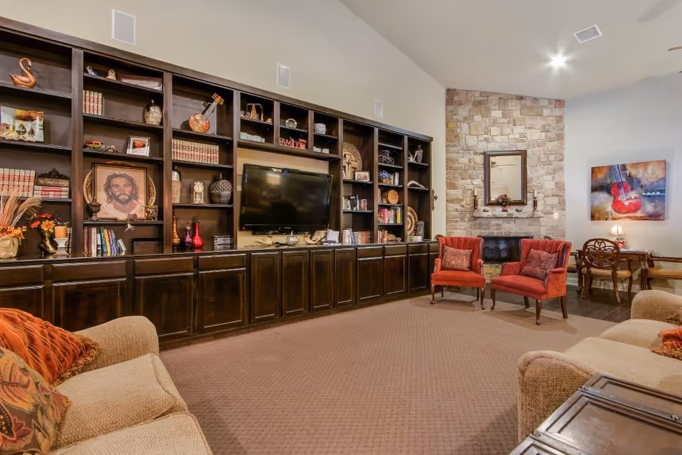 A cozy living room with beige sofas and orange cushions, two red armchairs, a large dark wooden built-in bookshelf with various decorative items and books, a flat-screen TV mounted in the center, and a stone fireplace with a mirror above it. There is also a small wooden desk with a chair and a colorful painting of a guitar on the wall.