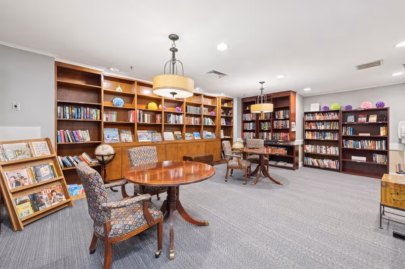 A well-lit library room with wooden bookshelves filled with books and decorative items. There are round wooden tables with patterned upholstered chairs arranged for reading or studying. The room has a carpeted floor and two ceiling lights with drum-shaped lampshades.