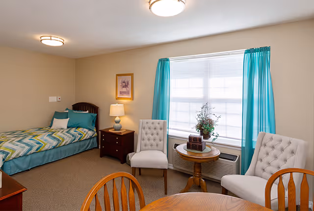 A cozy senior living room with a single bed dressed in teal and patterned bedding, a wooden nightstand with a lamp, two upholstered chairs, a round wooden table with decorative items, a large window with teal curtains, and a wooden dining table with chairs in the foreground.
