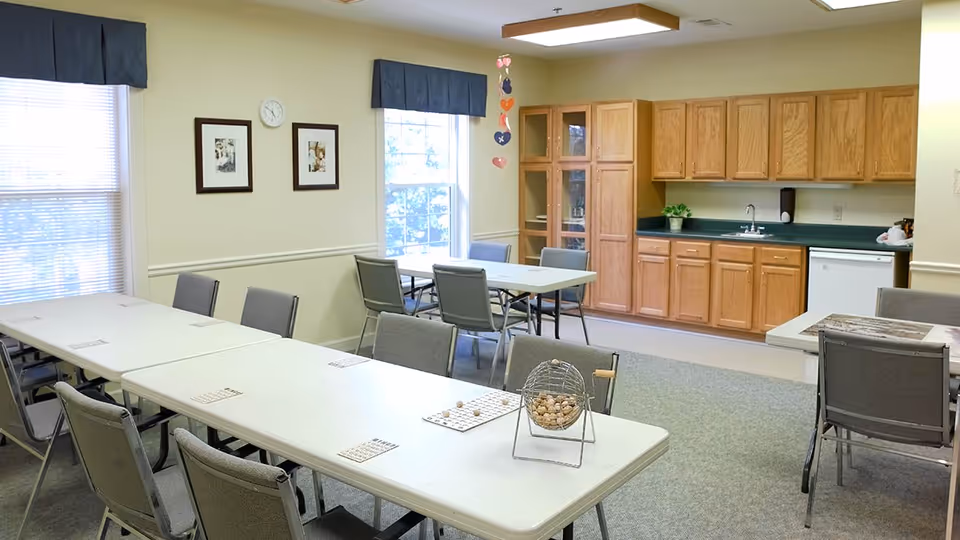 A bright room with multiple tables and chairs arranged for group activities. The tables have bingo cards and a bingo cage with balls. There are two large windows with blinds and valances, two framed pictures on the wall, and a clock. The back wall features wooden cabinets, a countertop with a sink, and a small refrigerator.