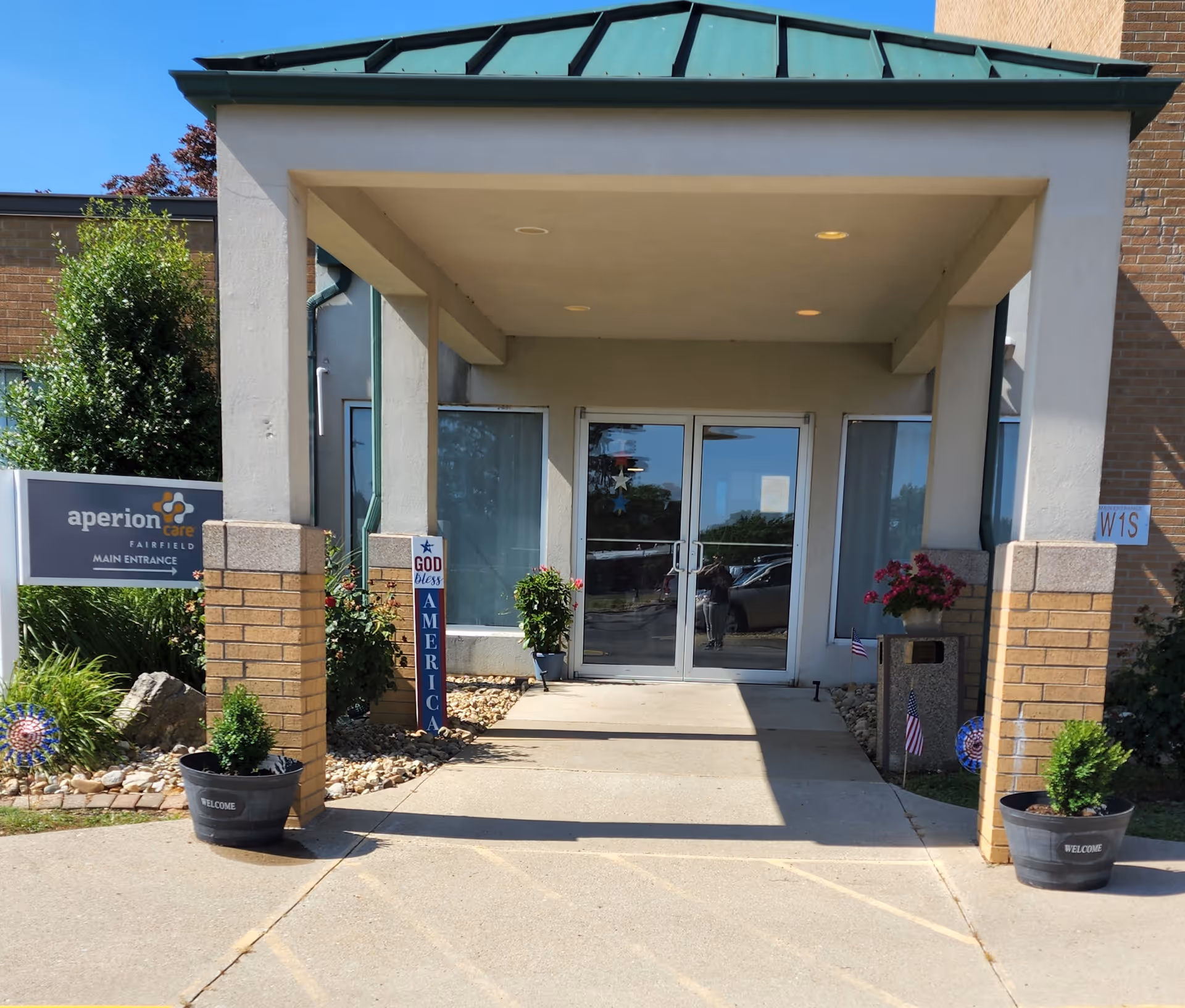 Main entrance of a senior living facility with a covered walkway supported by brick pillars. There are potted plants on either side of the entrance, a sign that reads 'aperion care FAIRFIELD MAIN ENTRANCE,' and a decorative sign that says 'God Bless America.' The entrance has double glass doors and windows on both sides.