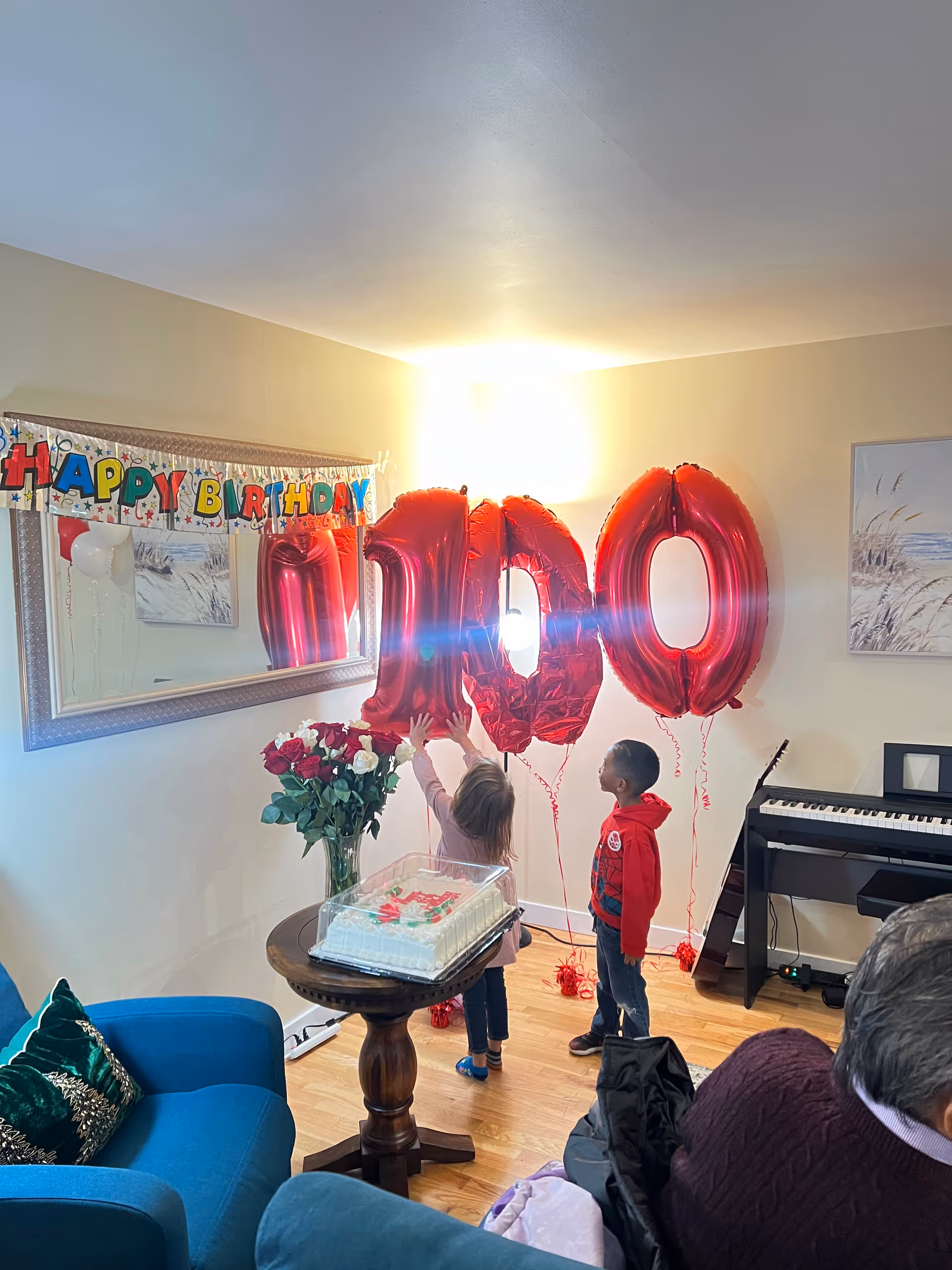 Two children in a living room reaching for large red "100" balloons beside a birthday cake, flowers, and a "Happy Birthday" banner.