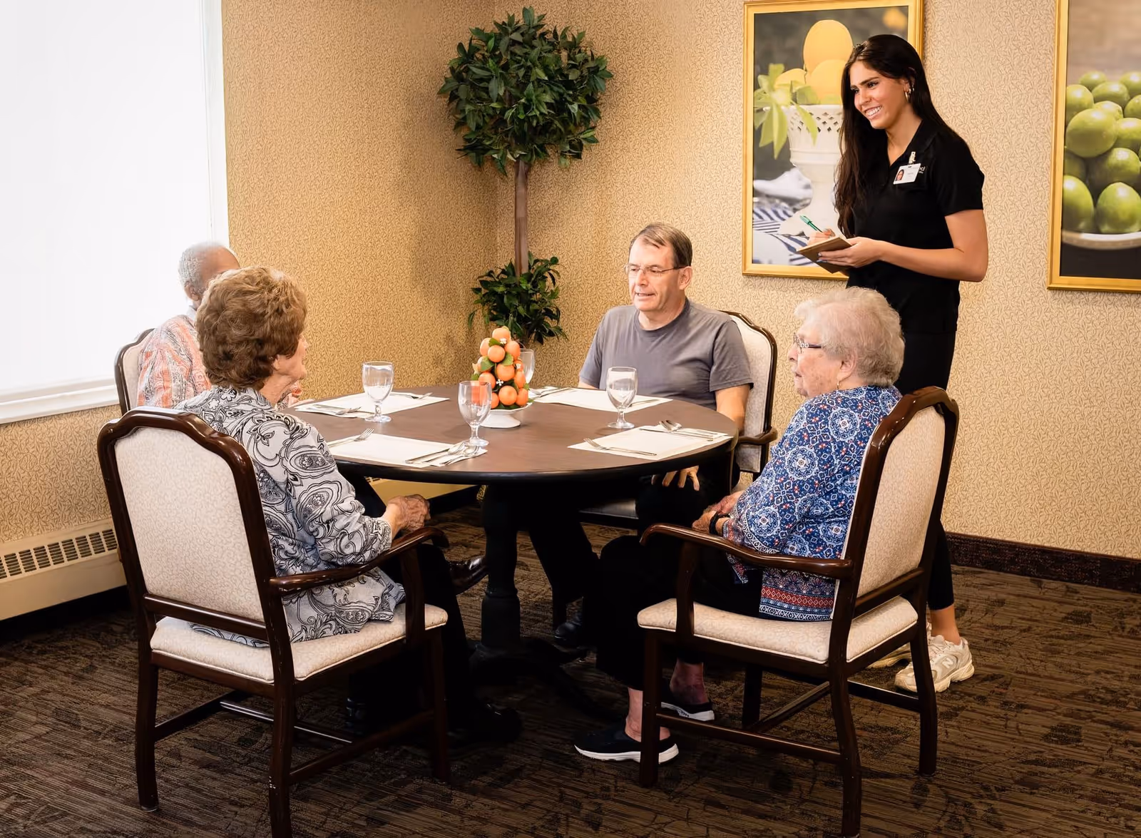 A group of four elderly people sitting around a round dining table set with glasses and napkins, while a young female staff member stands nearby holding a notepad and pen, in a warmly decorated room with framed fruit pictures on the wall and a potted plant in the corner.