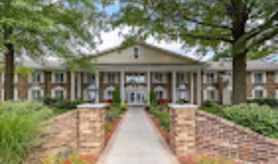 Front exterior view of a two-story senior living facility named Brown Deer Place, featuring a wide walkway flanked by brick pillars and landscaped greenery, with large trees and a blue sky in the background.