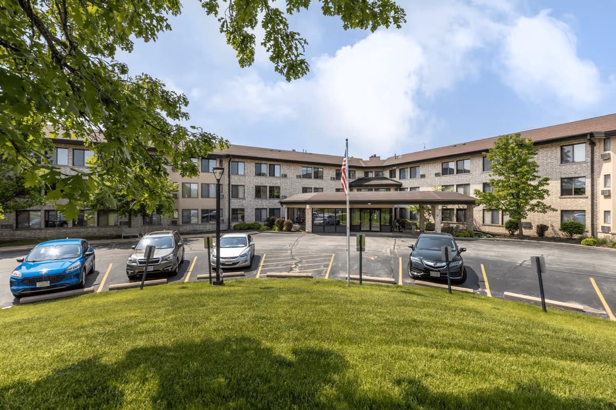 Front exterior view of Oak Hill Terrace Senior Living facility with a three-story building, a covered entrance, an American flag on a flagpole, several parked cars, green grass, and trees under a partly cloudy sky.