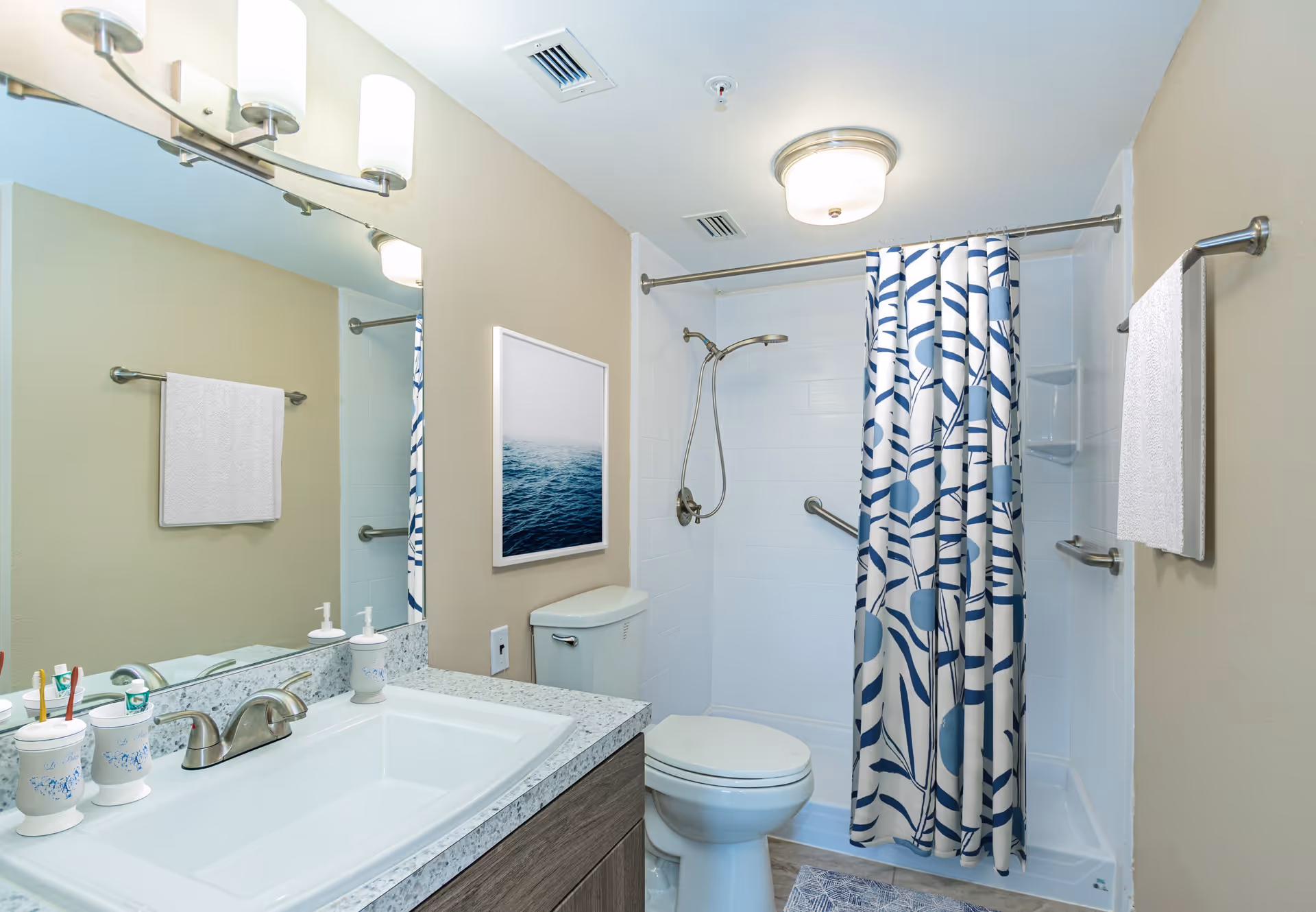 Clean accessible bathroom with sink and vanity, toilet, and a shower with a blue patterned curtain.