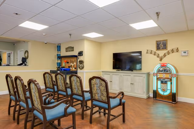 A common area with two rows of wooden chairs with blue cushions facing a flat-screen TV mounted on a white cabinet. To the right of the TV is a vintage jukebox with musical note wall decor above it. The room has beige walls, a wood floor, and a drop ceiling with fluorescent lights.