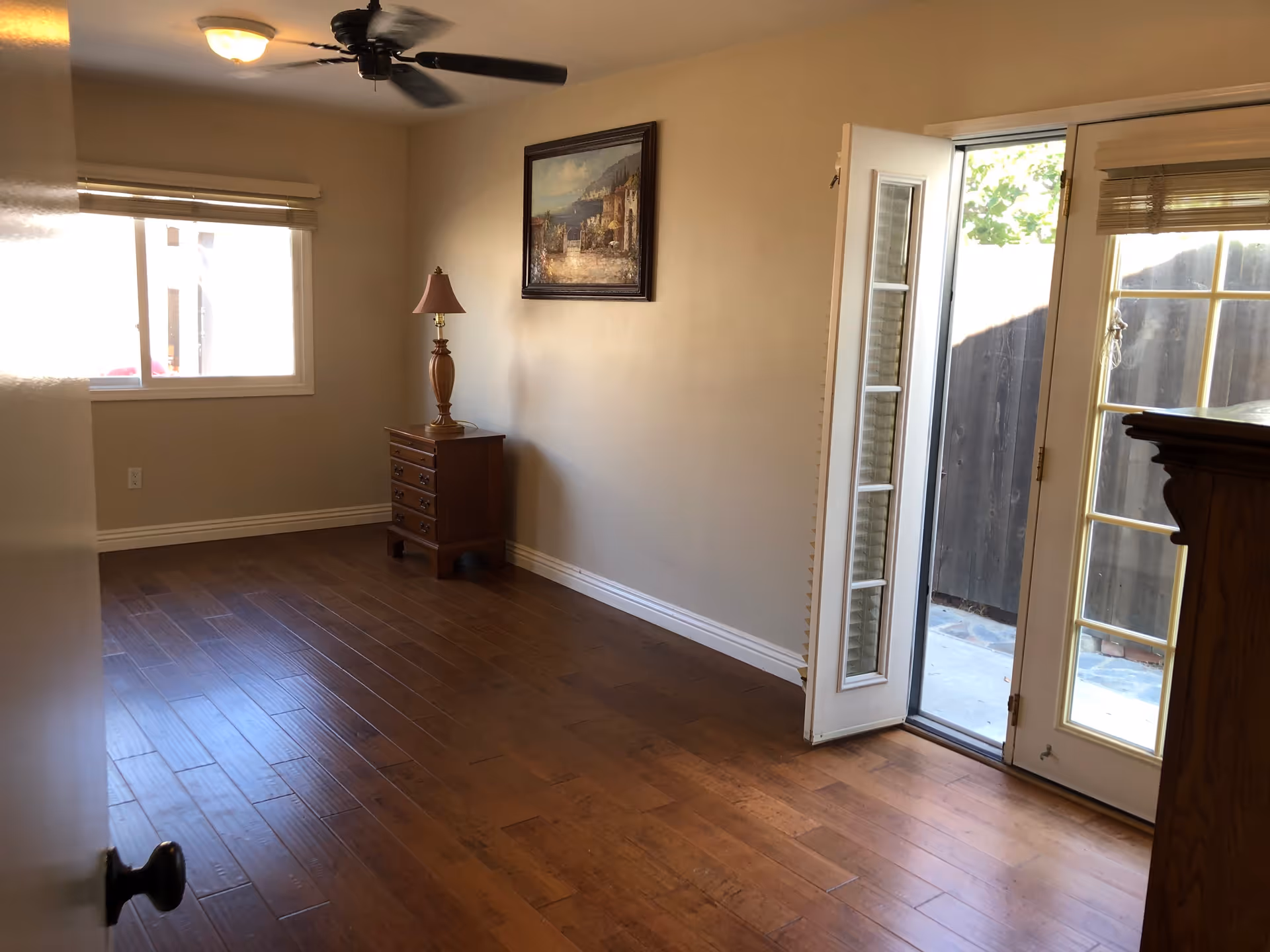 Empty room with wooden flooring, a small wooden chest of drawers with a lamp on top, a ceiling fan, a framed painting on the wall, a window with blinds, and a glass door leading outside to a fenced area.