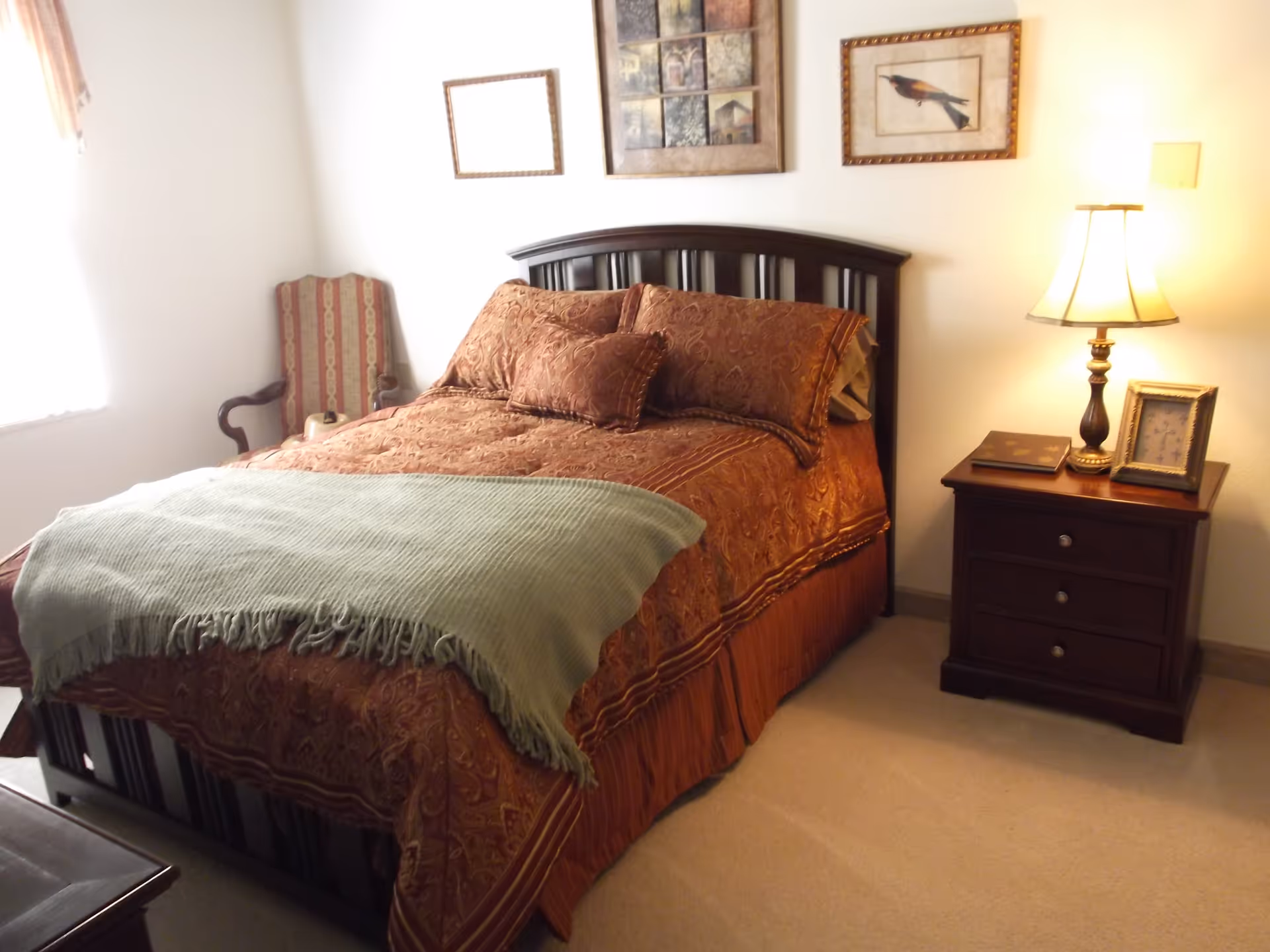 A warmly decorated bedroom with a made bed, decorative pillows, a nightstand with a lamp, and framed art on the wall.