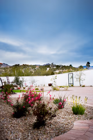 Outdoor garden area with a gravel bed containing various plants and flowers, a paved walkway, a white wall with a black gate, and a scenic view of hills and houses in the background under a partly cloudy sky.