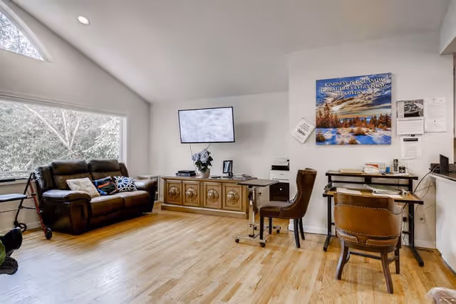 A bright living room with light wood flooring, a large window showing trees outside, a brown leather sofa with pillows, a walker beside it, a wall-mounted TV above a wooden cabinet, and two brown chairs at desks with office equipment and a scenic poster on the wall.