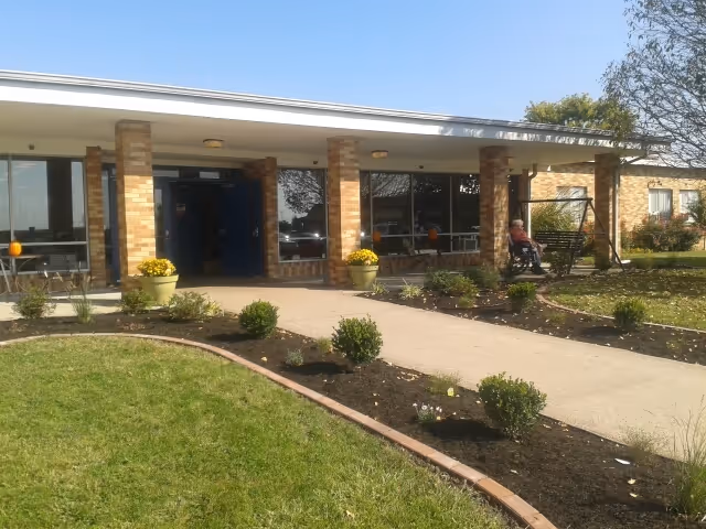 Exterior view of a single-story brick building with a covered entrance supported by brick columns. There is a paved walkway leading to the entrance, landscaped with small bushes and flowers. A person is sitting on a swing bench near the entrance. The sky is clear and blue.
