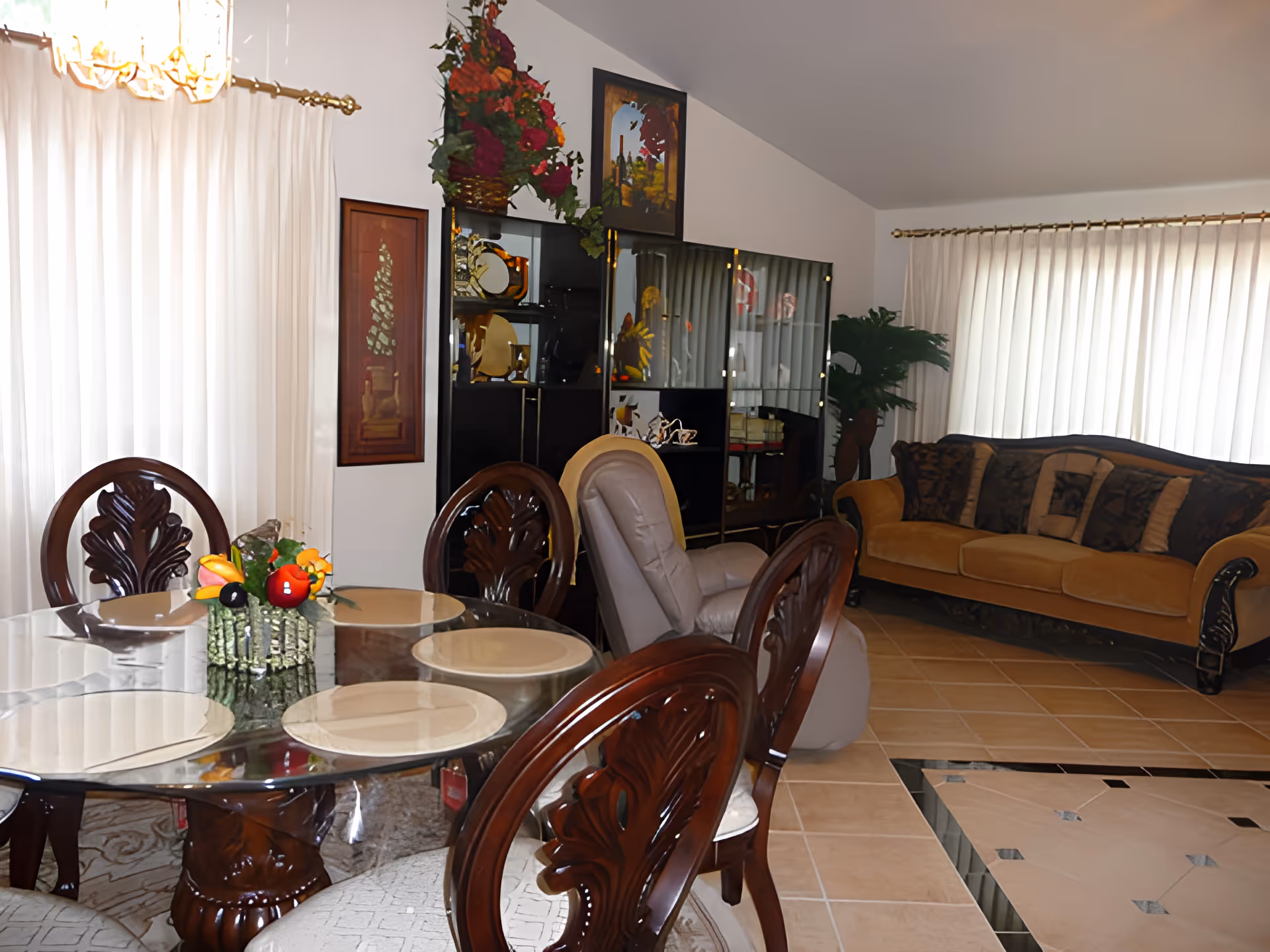 A cozy living and dining area featuring a glass-top dining table with four wooden chairs, a cushioned armchair, a brown sofa with patterned cushions, a black display cabinet with decorative items, and large windows with white curtains allowing natural light to fill the room.