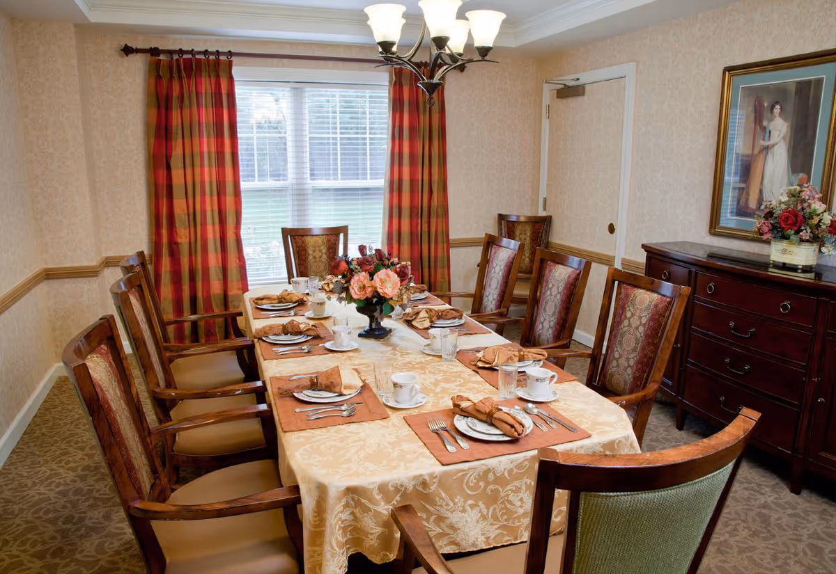 A formal dining room with a long table covered by a beige patterned tablecloth. The table is set with plates, cups, silverware, and brown napkins. A floral centerpiece is in the middle of the table. There are eight wooden chairs with upholstered seats and backs around the table. The room has patterned carpet, beige wallpaper, red and orange plaid curtains on a window, a chandelier overhead, a wooden sideboard with a flower arrangement, and a framed painting on the wall.