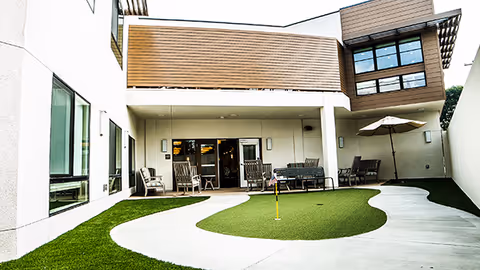 Outdoor courtyard area at Avenir Memory Care Westside featuring a putting green with a small flag, surrounded by a curved concrete walkway. There are several chairs and tables with umbrellas along the building walls, and the building has large windows and a modern design.