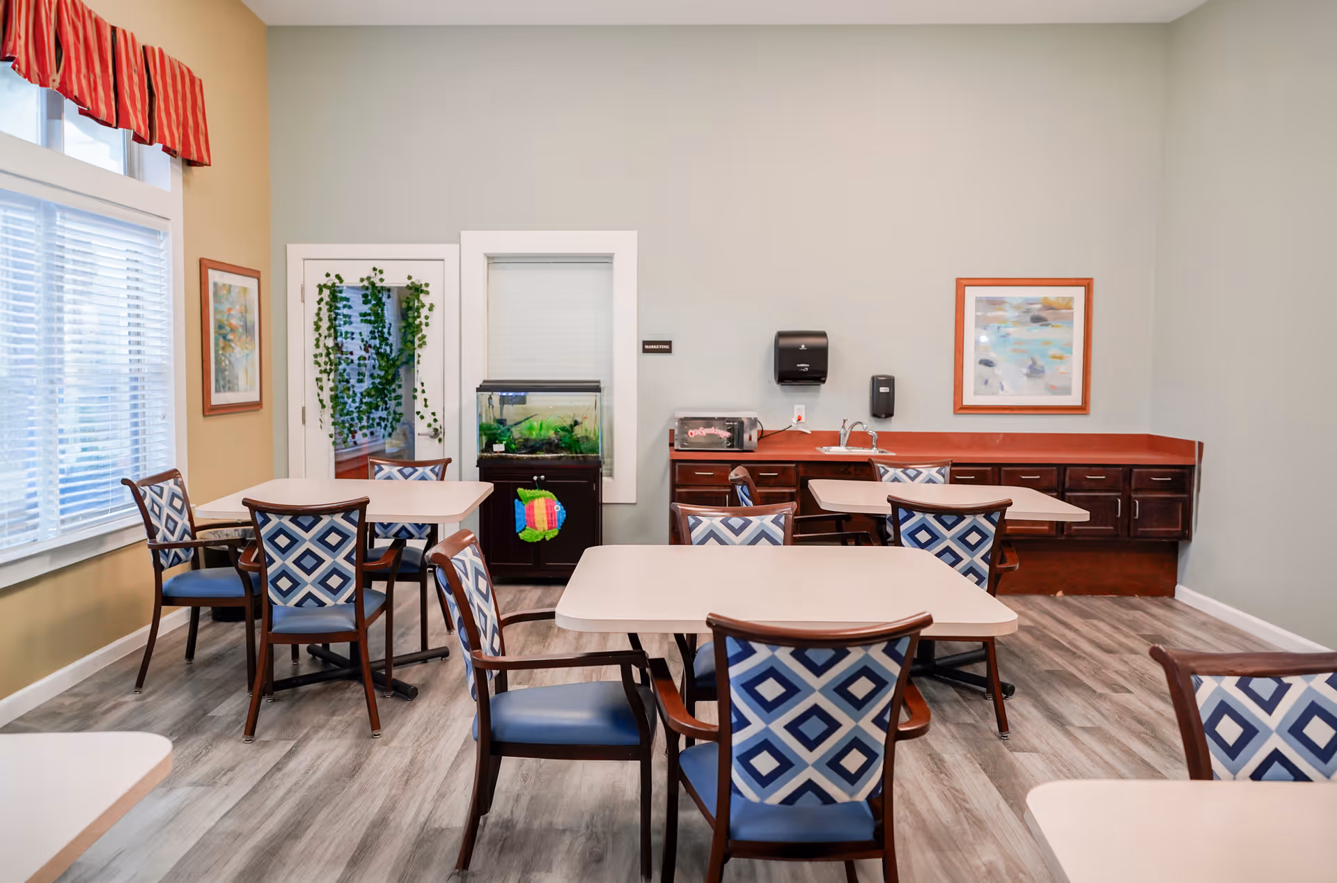 A bright dining area in a senior living facility with several tables and chairs featuring blue and white patterned upholstery. The room has large windows with blinds and red valances, light-colored walls, and wood-look flooring. There is a small fish tank on a stand, a countertop with a sink, a paper towel dispenser, and framed artwork on the walls.