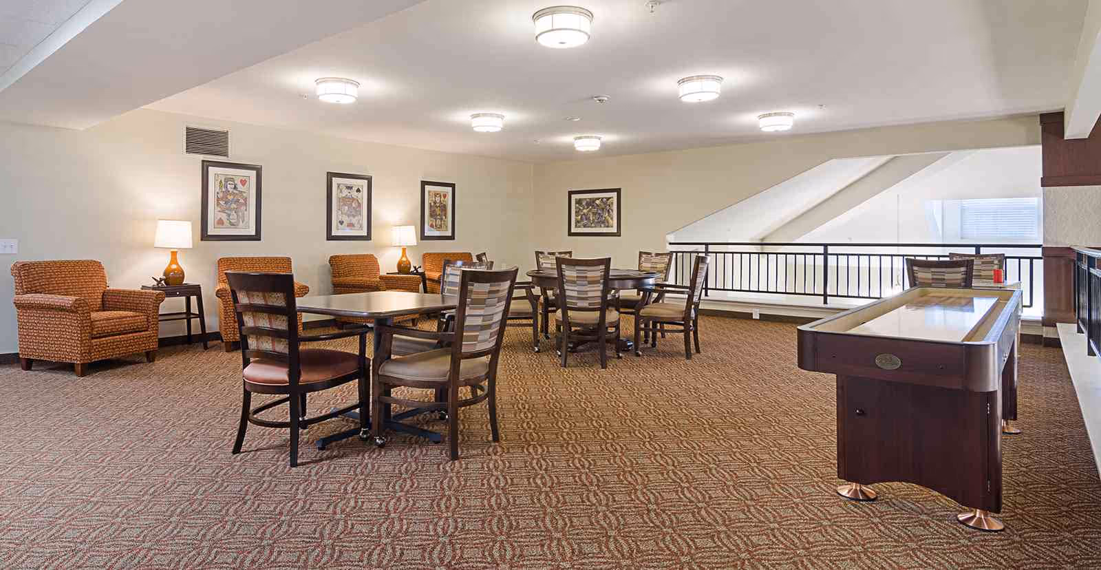 A spacious common area with patterned carpet, several tables and chairs arranged for seating, a few armchairs with side tables and lamps, framed artwork on the walls, and a shuffleboard table near a railing overlooking a lower level.