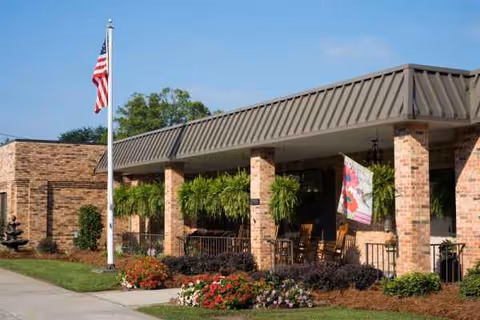Exterior view of a brick senior living facility with a covered porch, flagpole, and seating.