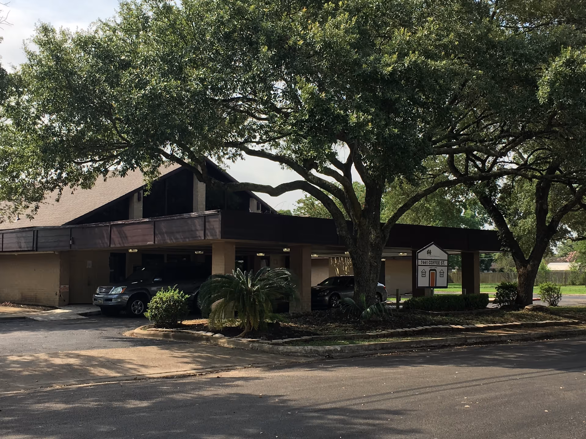 Exterior view of a single-story building with a sloped roof partially shaded by large trees. There are two cars parked under a covered area near the entrance. A sign near the entrance displays the address 7441 Coffee St.