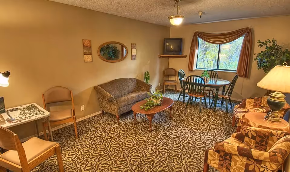 A cozy living room area in a senior living facility with patterned carpet, a small couch, a wooden coffee table with a plant, a round dining table with four chairs near a window with brown curtains, two patterned armchairs with a side table and lamp, a small table with chairs and a puzzle on it, and a TV mounted on a corner shelf.