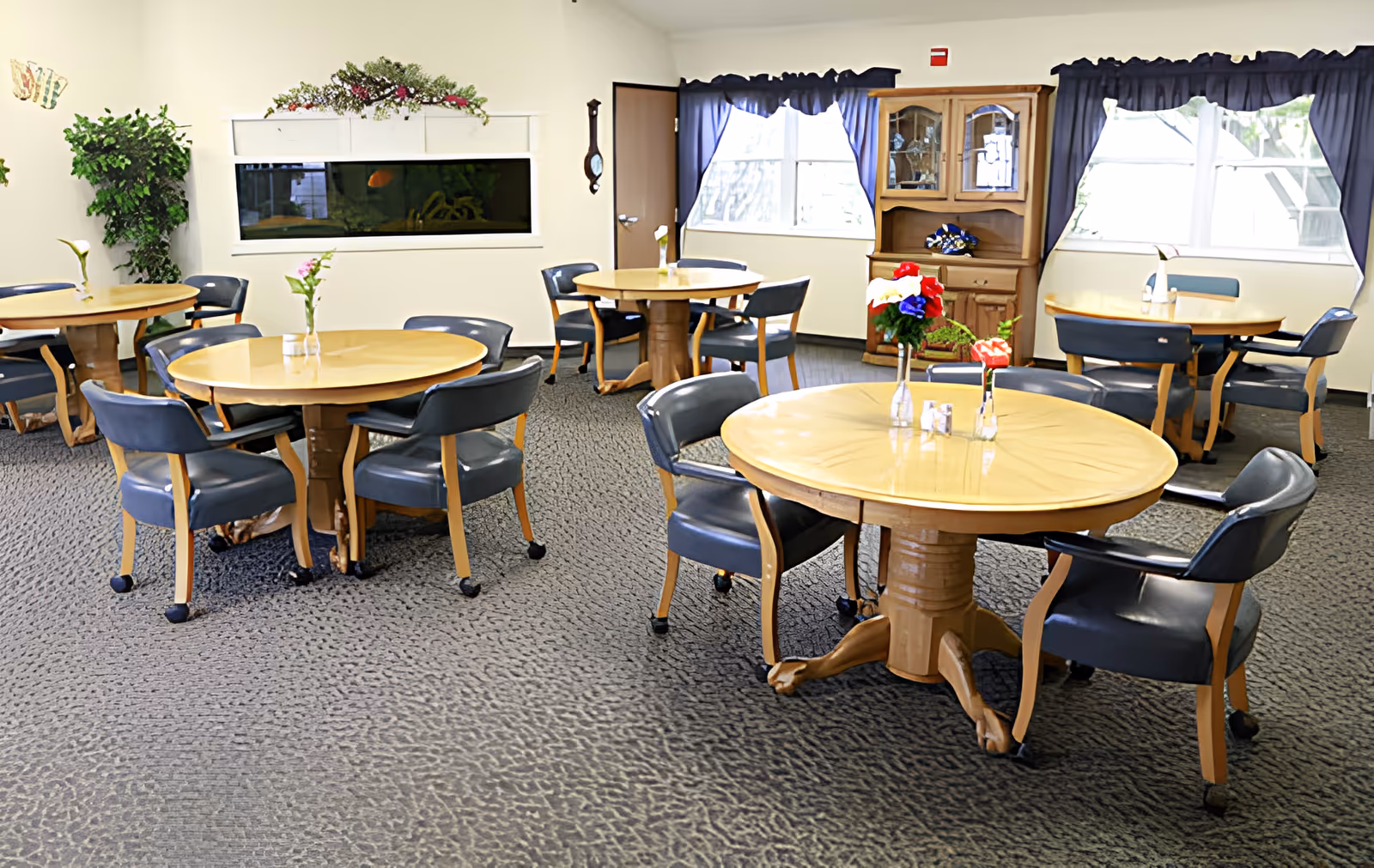 Communal dining room with round wooden tables, navy cushioned chairs, small vases of flowers, windows and an aquarium.
