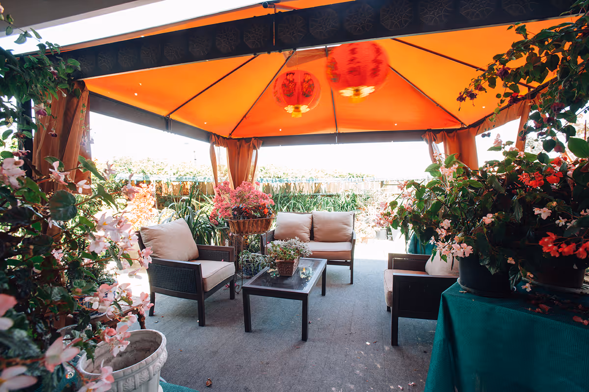 Covered outdoor seating area with wicker chairs and a sofa under an orange canopy surrounded by potted flowering plants.