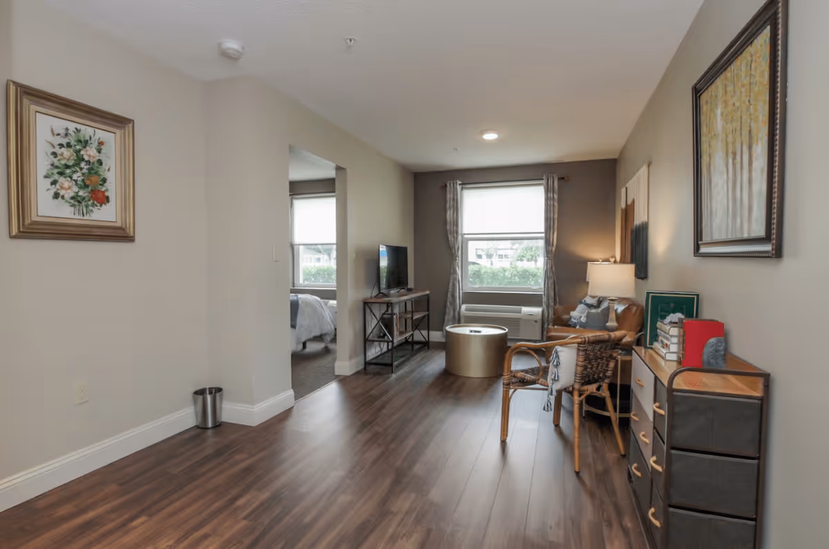 A cozy senior living room with wooden flooring, a wicker chair with a cushion, a small round coffee table, a TV on a stand, and a window with curtains letting in natural light. A dresser with books and a lamp is on the right side, and a bedroom is visible through an open doorway.