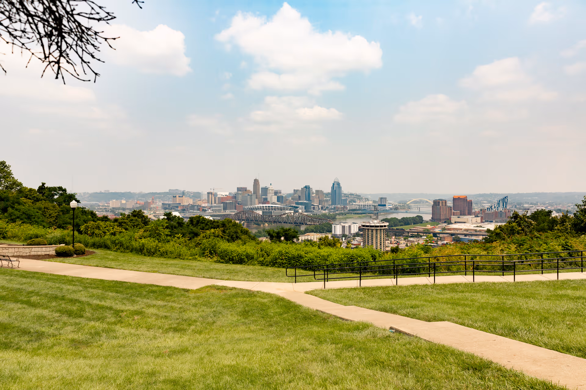A scenic outdoor view from a grassy hill overlooking a city skyline in the distance under a partly cloudy sky. There are paved walkways, green bushes, and trees in the foreground.