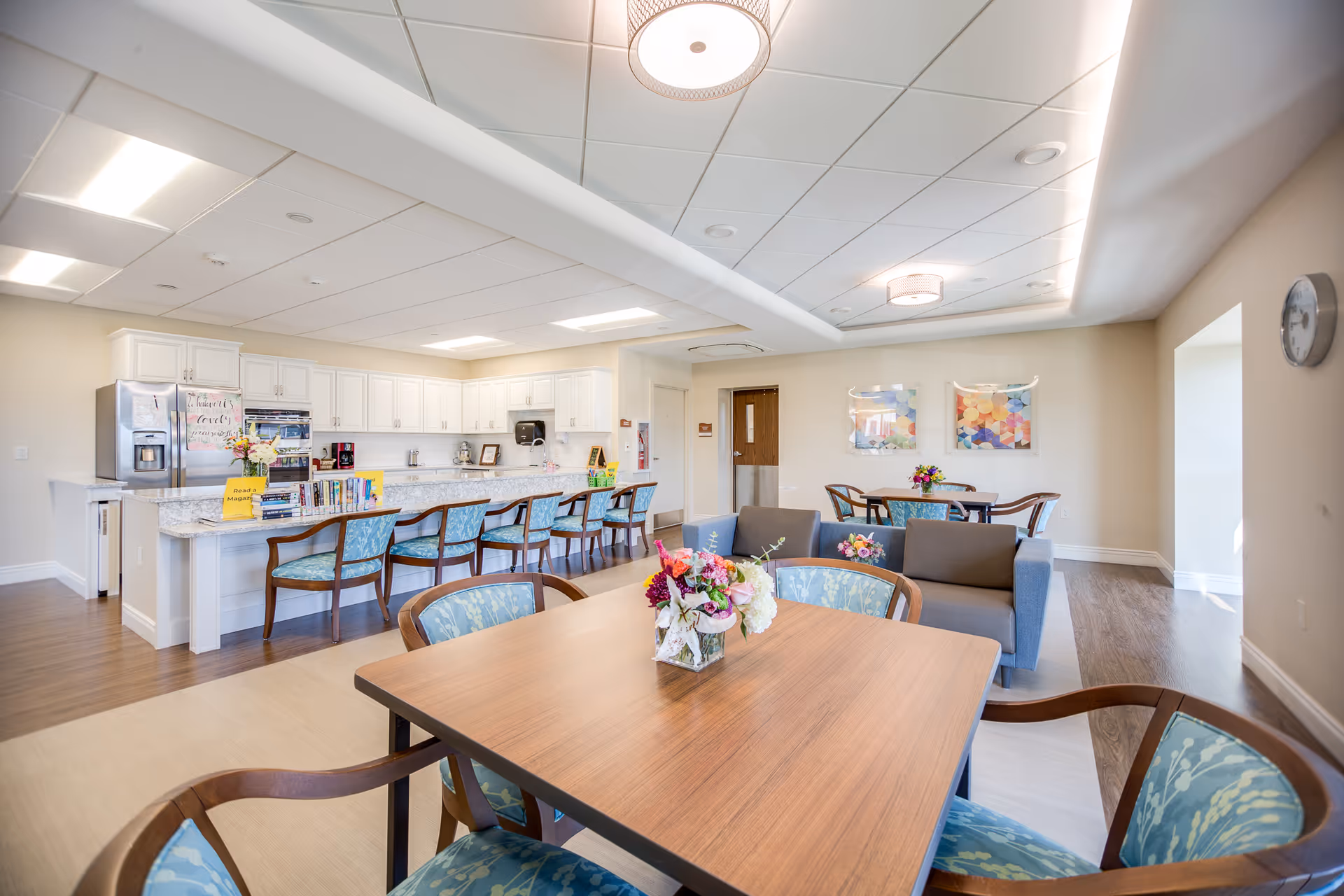 A bright and spacious common area featuring a modern kitchen with white cabinets, a long granite countertop with several chairs, and a dining area with wooden tables and chairs. The room is decorated with colorful flower arrangements on the tables and has a light, neutral color palette with wood flooring and a white ceiling with recessed lighting.