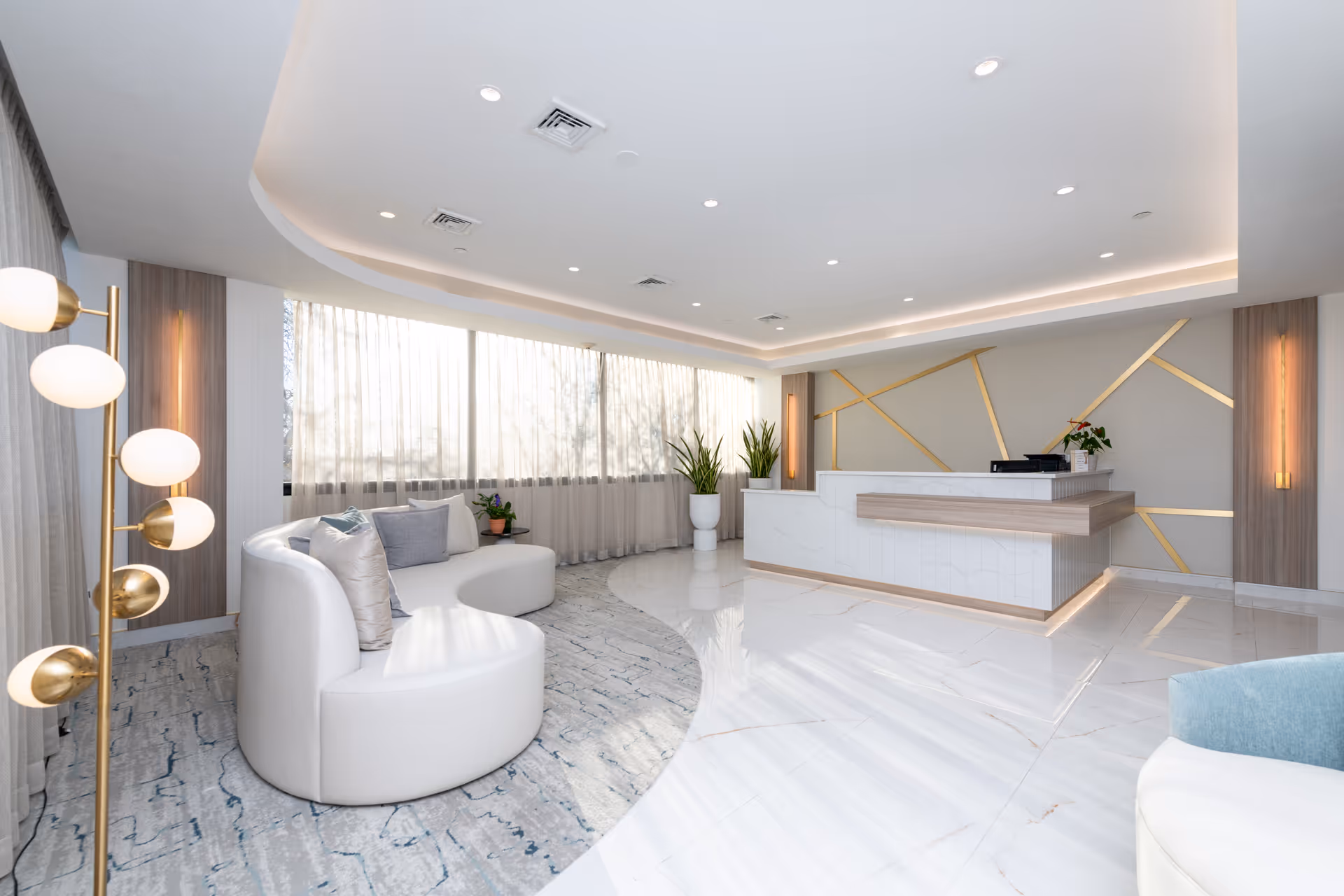 A modern, bright reception area with a curved white sofa adorned with cushions, a stylish floor lamp with round white bulbs, large windows with sheer curtains allowing natural light, and a reception desk with geometric gold accents on the wall behind it. There are also potted plants adding greenery to the space.
