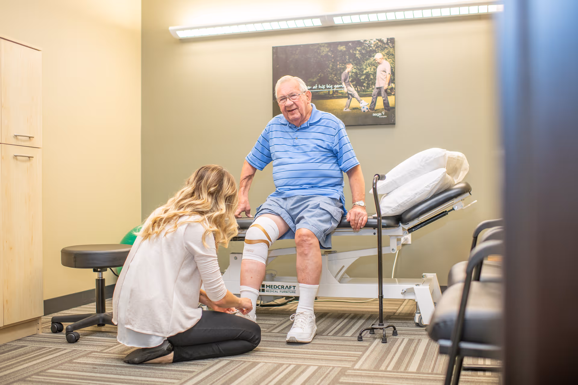An elderly man sitting on a medical examination table with a knee brace, while a woman kneels in front of him adjusting the brace in a clinical room with beige walls and a framed picture on the wall.