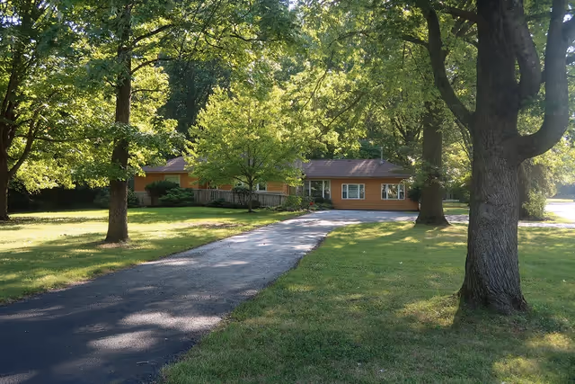 A single-story building with a brown exterior surrounded by large green trees and a well-maintained grassy lawn. A paved driveway leads up to the building, which is partially shaded by the trees.
