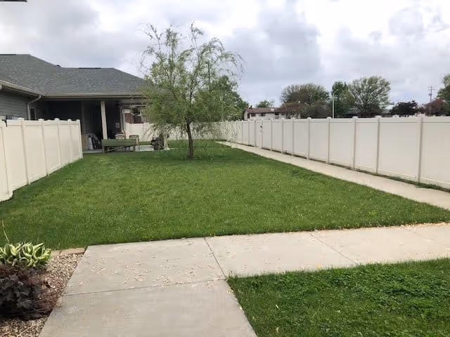 A fenced outdoor area with green grass, a small tree in the center, and a concrete walkway running alongside the grass. There is a building with a covered porch on the left side under a cloudy sky.