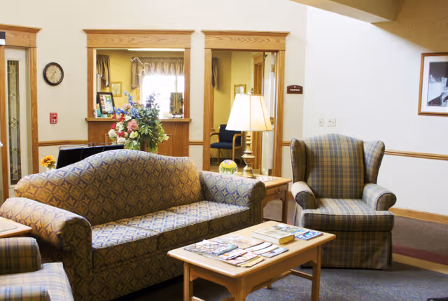 Communal living room with a patterned sofa and armchairs around a coffee table with magazines, a lamp, and a floral arrangement near a reception window.