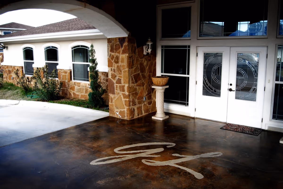 Entrance area of a facility with a covered driveway, stone walls, white double doors with decorative glass panels, and a concrete floor with the initials 'GL' painted on it. There are windows along the side of the building and some plants near the wall.