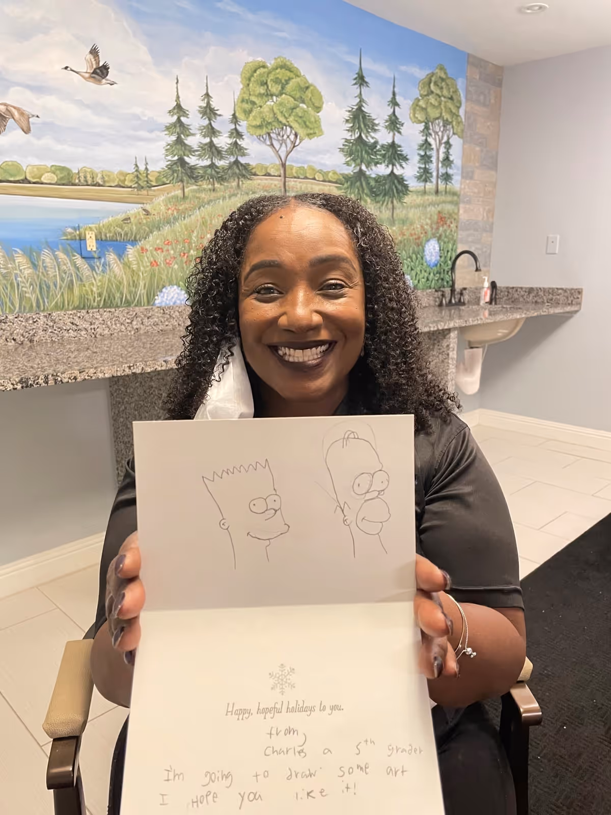 A smiling woman with curly hair is seated indoors, holding up a card with pencil drawings of Bart and Homer Simpson. Behind her is a wall mural depicting a nature scene with trees, grass, and birds near a body of water. The room has a granite countertop with a sink and light-colored tiled floor.