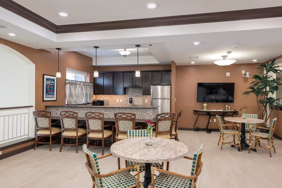 Interior view of a communal dining and kitchen area with a counter and six chairs, two round tables with patterned chairs, a wall-mounted TV, and a large potted plant. The walls are painted brown and the floor is tiled.